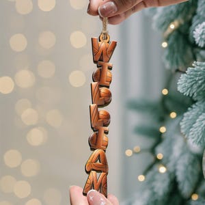 May include: Wooden ornament spelling out the name "William" with basketballs incorporated into the letters. The ornament is brown and has a twine loop for hanging. The background is blurred with Christmas tree and lights.