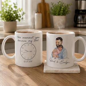May include: Two white ceramic mugs with coffee. One mug has a clock design and the text "The moment you became my Dad" and "02.01.2026". The other mug has a drawing of a father holding a baby and the text "Dad & Evelyn".