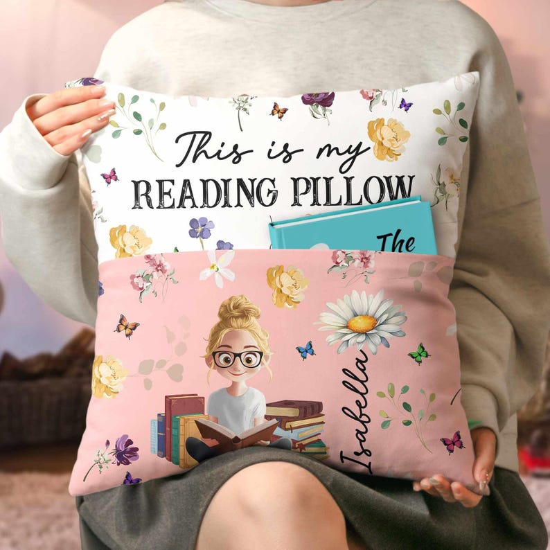 May include: A reading pillow with a pocket, decorated with flowers and butterflies, and the text "This is my READING PILLOW." The front of the pillow is pink, with a cartoon girl reading, the name "Isabella," and a book.