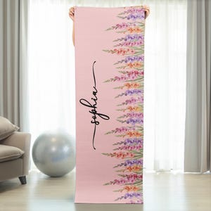 May include: A rectangular yoga mat in a soft pink hue featuring a vertical arrangement of watercolor-style flowers in shades of purple, orange, and pink along the right side. The name Sophia is printed in a black cursive script on the left side.