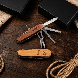 May include: Two personalised wooden multi-tools with various tools extended, including a knife, saw, and scissors. One tool is engraved with "MKS" and the other with "JIS". The tools are displayed on a dark wooden surface.