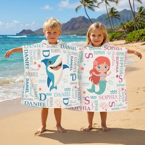May include: Two children on a beach holding personalized hooded towels. One towel features a blue shark graphic with the name "Daniel" repeated. The other towel has a mermaid graphic with the name "Sophia".
