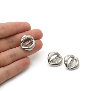 Könnte beinhalten: Silberfarbene Ohrringe mit einem einzigartigen, offenen Design. Jeder Ohrring hat eine geschwungene, ovale Form mit einer Reihe kleiner, klarer Steine in der Mitte. Die Ohrringe werden auf weißem Hintergrund präsentiert.