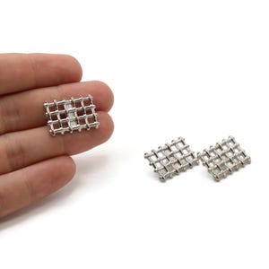May include: Silver-toned metal cufflinks with a grid design. One cufflink is held in a hand, while two others are displayed on a white background. The cufflinks have a rectangular shape with a woven pattern.