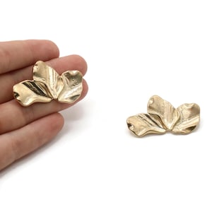 May include: Two gold-colored, abstract flower-shaped jewelry pieces. One is held in a hand, the other is separate. The pieces have a textured, folded petal design and are set against a white background.