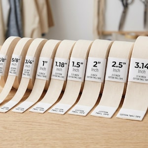 Op de afbeelding: Een verzameling beige katoenen keperbandrollen in verschillende breedtes, van 1 cm tot 8 cm. Elke rol is gelabeld met de maat in inches en de woorden "Cotton Twill Tape". De rollen worden getoond op een licht houten oppervlak.