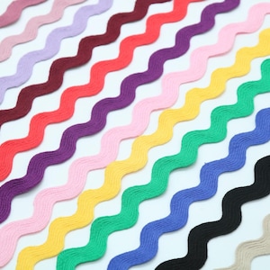 Puede incluir: Una colección de cintas onduladas multicolores en varios tonos, incluyendo rosa, morado, rojo, amarillo, verde, azul, negro y beige. Las cintas están dispuestas diagonalmente, creando una exhibición vibrante y texturizada, ideal para manualidades.