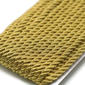 May include: Close-up of a length of gold-colored twisted rope trim. The trim has a textured, braided appearance and is likely used for decorative purposes in crafts or sewing projects. The rope is lying on a white surface.