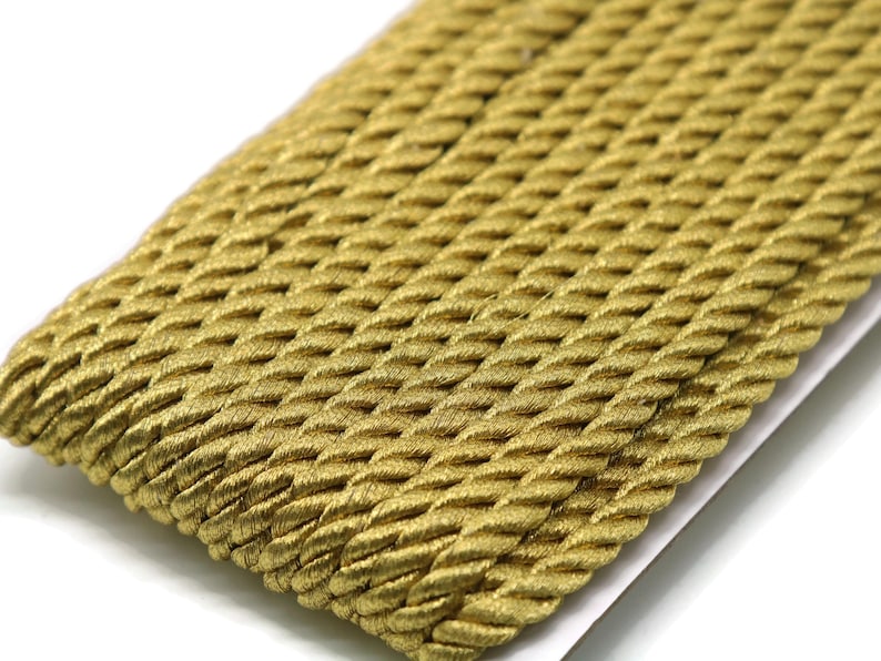 May include: Close-up of a length of gold-colored twisted rope trim. The trim has a textured, braided appearance and is likely used for decorative purposes in crafts or sewing projects. The rope is lying on a white surface.