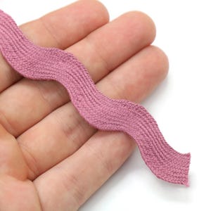 May include: A length of mauve-colored, wavy ribbon. The ribbon has a textured, woven appearance and is held in a hand against a white background. The ribbon's edges are slightly frayed.