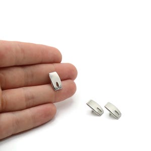 Puede incluir: Pendientes de metal plateado con forma rectangular curvada y un pequeño recorte. Un pendiente se sostiene en una mano, mientras que otros dos se exhiben sobre una superficie blanca. Diseño minimalista.