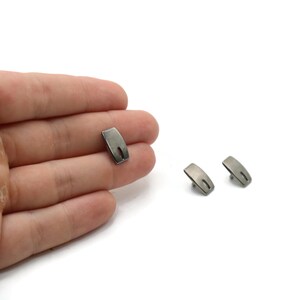 May include: Three small, metallic gray objects with a curved rectangular shape and a small, oval cutout. One object is held in a hand, while the other two are on a white surface. The objects appear to be hardware components.