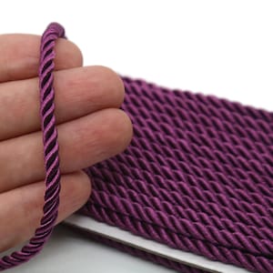 May include: Close-up of a length of twisted purple cord. The cord is held in a hand, with a larger quantity of the same cord visible in the background. The cord has a twisted design and a glossy finish, suitable for crafts and decorative projects.
