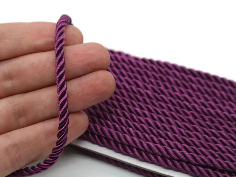May include: Close-up of a length of twisted purple cord. The cord is held in a hand, with a larger quantity of the same cord visible in the background. The cord has a twisted design and a glossy finish, suitable for crafts and decorative projects.