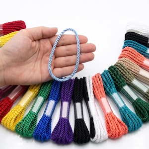 May include: A variety of colorful twisted cords, including red, yellow, green, blue, purple, black, white, and coral. A light blue cord is held in a hand. The cords are bundled and wrapped with clear plastic.