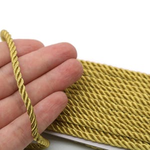 May include: Close-up of a golden-yellow twisted rope. The rope is held in a hand, with a larger pile of the same rope visible in the background. The rope has a shiny, textured appearance, suggesting it may be made of a satin or similar material. The background is white.