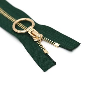 May include: A close-up of a dark green zipper with a gold-colored pull and teeth. The zipper is open, revealing the teeth and the fabric tape. The pull has a circular ring.