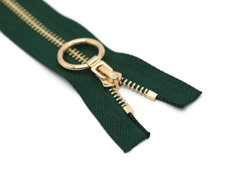 May include: A close-up of a dark green zipper with a gold-colored pull and teeth. The zipper is open, revealing the teeth and the fabric tape. The pull has a circular ring.