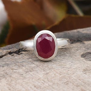 May include: A silver ring featuring an oval-cut, deep red ruby gemstone. The ruby is set in a bezel setting, and the band is a simple, polished silver. The ring is displayed on a wooden surface, with blurred foliage in the background.