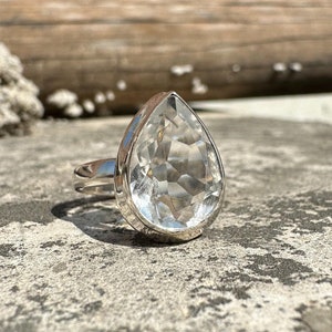 Op de afbeelding: Een zilveren ring met een heldere, traanvormige kwarts edelsteen. De ring ligt op een grijs, getextureerd oppervlak.