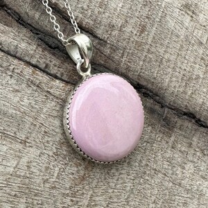 May include: A silver necklace with a round, pale pink pendant. The pendant features a light pink stone set in a silver bezel with a decorative edge. The chain is a delicate silver link.