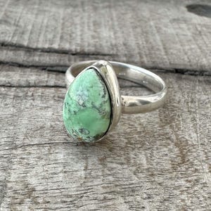 May include: A silver ring featuring a pear-shaped, light green turquoise stone. The stone has a mottled pattern with darker green and brown inclusions. The ring band is a simple, polished silver.