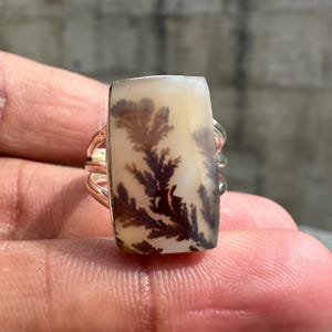 May include: A rectangular dendritic agate gemstone set in a polished metal ring. The stone features a translucent, creamy white base with dark brown, fern-like inclusions that branch across the surface, held by a multi-banded metal setting.