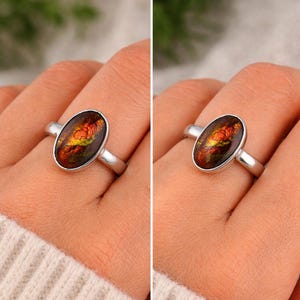May include: A silver ring featuring an oval-shaped ammolite gemstone with iridescent flashes of red, orange, and green. The stone is set in a smooth, polished silver bezel on a simple, slender silver band that encircles the finger.