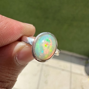 Può includere: Anello in argento con un'opale ovale. L'opale mostra un vibrante gioco di colori, con lampi di verde, giallo e rosso su una base bianco latte. La fascia dell'anello è in argento lucido.
