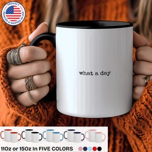 May include: White ceramic mug with a black rim and handle. The mug has the text "what a day" printed in black on the front. The mug is available in five colors: red, blue, black, white, and pink.