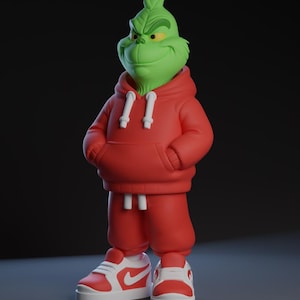 Op de afbeelding: Een groene Grinch-figuur met een rode hoodie, bijpassende joggingbroek en rood-witte sneakers. De Grinch heeft een ondeugende uitdrukking en poseert met zijn handen in de zakken van de hoodie. De figuur staat tegen een donkere achtergrond.