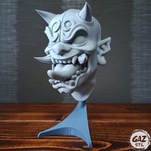 May include: A grey 3D-printed Oni mask sculpture, a Japanese demon with horns, sharp teeth, and a protruding tongue. The mask is mounted on a blue stand. The "GAZ STL" logo is visible on the wooden surface.