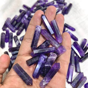 May include: A collection of amethyst crystal points, also known as wands, in various shades of purple. The crystals are smooth and polished, with a pointed tip and a flat base. The crystals are arranged in a pile, with some overlapping.