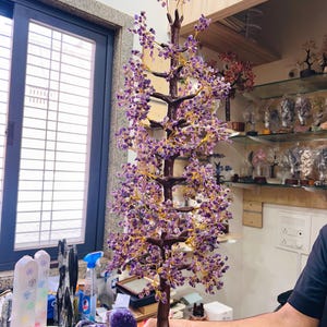 以下が含まれることがあります： アメジストの装飾的な木の彫刻。木は、紫のアメジストの結晶のクラスターで飾られた濃い茶色の幹と枝が特徴です。結晶は金色のワイヤーで取り付けられています。木は、粗い天然アメジストのベースに取り付けられています。