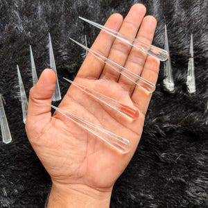 Puede incluir: Una colección de puntas de cristal de cuarzo transparente, también conocidas como varitas, que se muestran en una mano. Las puntas son largas y delgadas, con una punta afilada. Son una opción popular para la curación con cristales y la meditación.