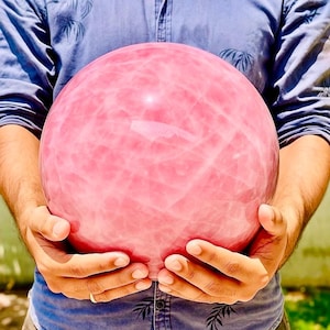 Op de afbeelding: Een grote, gladde, bolvormige rozenkwarts met een doorschijnende, lichtroze tint en zichtbare interne breuken en troebele witte insluitingen over het gehele gepolijste oppervlak, stevig vastgehouden in beide handen.