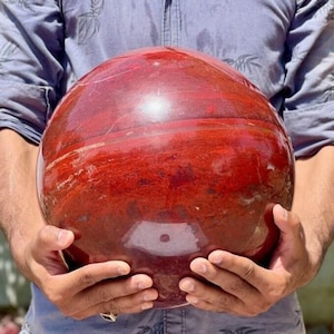 Puede incluir: Una gran esfera roja pulida hecha de piedra natural. La esfera tiene un acabado liso y brillante y un color rojo intenso con sutiles variaciones de tono.