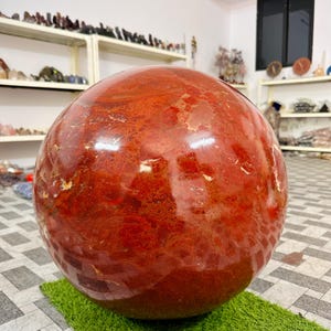 Könnte beinhalten: Ein großer, polierter kugelförmiger Stein in tiefem Rot mit komplexen natürlichen Mustern aus dunkleren karmesinroten und helleren orangebraunen Einschlüssen auf der glatten, glänzenden Oberfläche, der massiv und schwer wirkt.