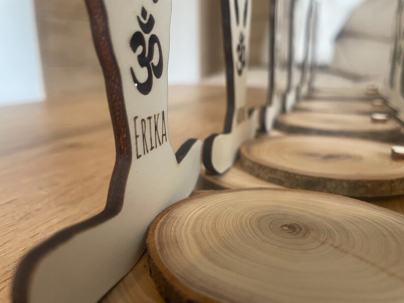 K&ouml;nnte beinhalten: Ein Satz Holzfiguren in Yoga-Posen mit dem Om-Symbol und Namen auf jeder Figur. Die Figuren stehen auf Holzscheiben.