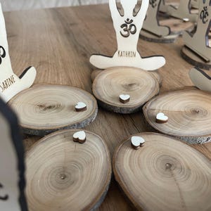 K&ouml;nnte beinhalten: F&uuml;nf Holz-Untersetzer mit einem Yoga-Motiv und den Namen Sabine, Kathrin und Erika. Jeder Untersetzer hat ein kleines wei&szlig;es Herz oben drauf.