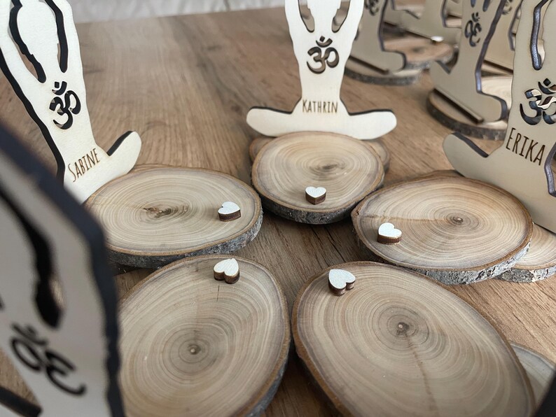 K&ouml;nnte beinhalten: F&uuml;nf Holz-Untersetzer mit einem Yoga-Motiv und den Namen Sabine, Kathrin und Erika. Jeder Untersetzer hat ein kleines wei&szlig;es Herz oben drauf.