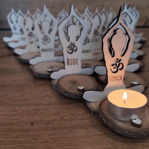 K&ouml;nnte beinhalten: Eine Reihe von Kerzenhaltern aus Holz in Form einer Person in einer Yoga-Pose. Jeder Halter hat ein Om-Symbol und einen Namen, wie "Rudi" und "Sonja". Eine brennende Teelichtkerze befindet sich in einem der Halter.
