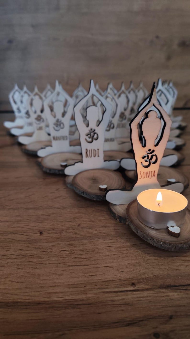 K&ouml;nnte beinhalten: Eine Reihe von Kerzenhaltern aus Holz in Form einer Person in einer Yoga-Pose. Jeder Halter hat ein Om-Symbol und einen Namen, wie "Rudi" und "Sonja". Eine brennende Teelichtkerze befindet sich in einem der Halter.