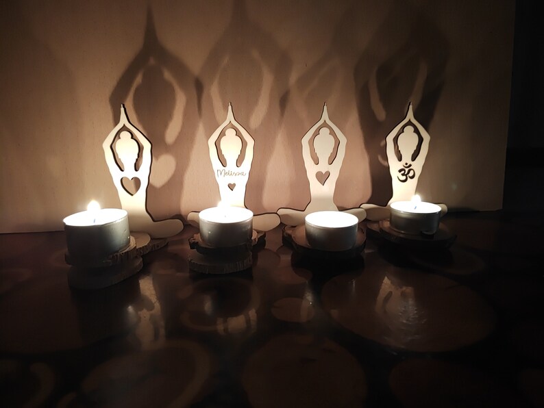 K&ouml;nnte beinhalten: Vier Kerzenhalter aus Holz in Form von Yoga-Posen, jeder mit einer brennenden Teelichtkerze. Die Halter sind in einer Reihe auf einer dunklen Oberfl&auml;che angeordnet. Der mittlere Halter tr&auml;gt die Aufschrift "Welcome".