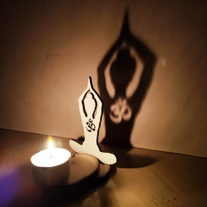 K&ouml;nnte beinhalten: Eine Holz-Ausschneidung einer Person in einer Yoga-Pose mit dem Om-Symbol auf der Brust, die einen Schatten an eine Wand wirft. Eine kleine Kerze brennt vor der Ausschneidung.