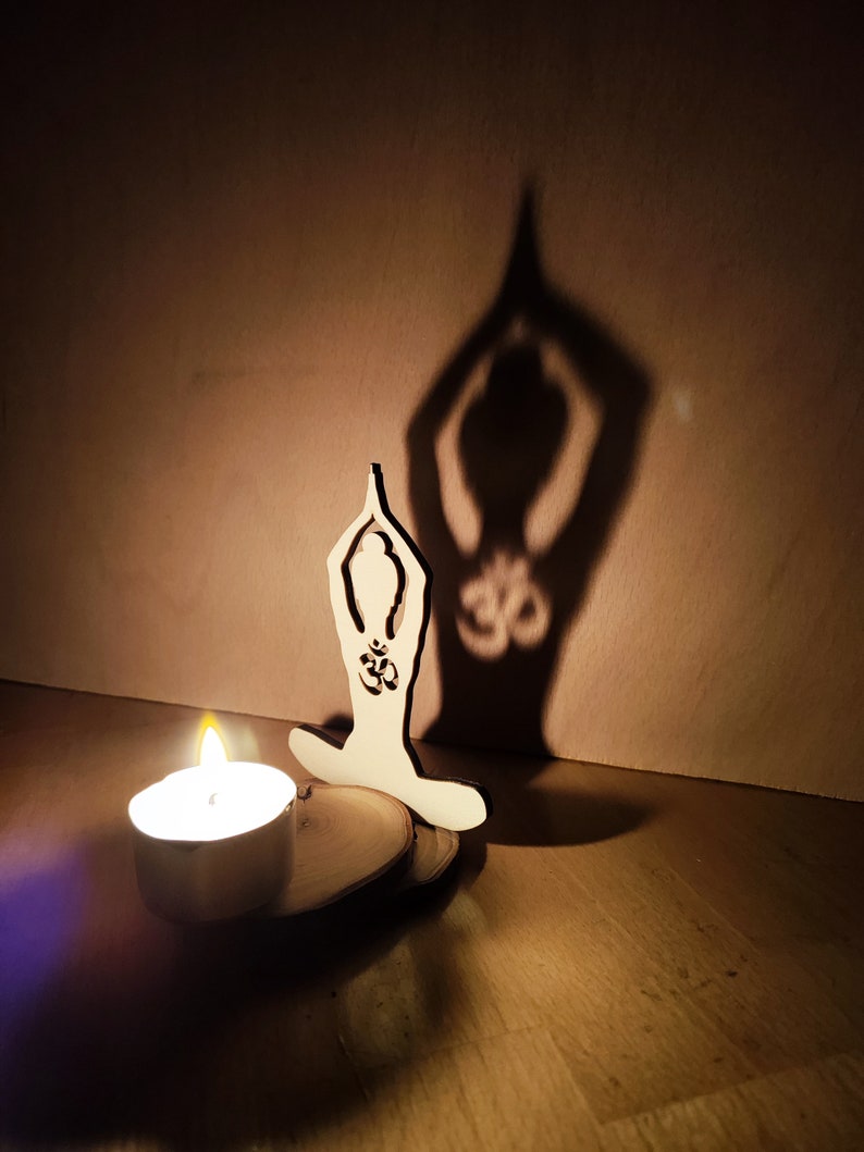K&ouml;nnte beinhalten: Eine Holz-Ausschneidung einer Person in einer Yoga-Pose mit dem Om-Symbol auf der Brust, die einen Schatten an eine Wand wirft. Eine kleine Kerze brennt vor der Ausschneidung.