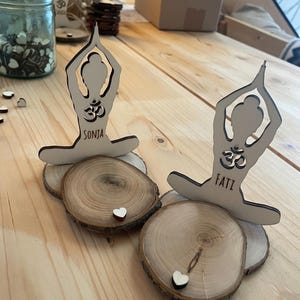 K&ouml;nnte beinhalten: Zwei Holzfiguren in Yoga-Pose mit dem Wort "Om" und den Namen "Sonja" und "Fati" darauf. Die Figuren stehen auf Holzscheiben mit kleinen wei&szlig;en Herzen oben drauf.