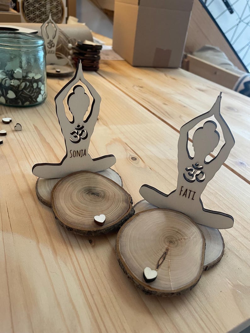 K&ouml;nnte beinhalten: Zwei Holzfiguren in Yoga-Pose mit dem Wort "Om" und den Namen "Sonja" und "Fati" darauf. Die Figuren stehen auf Holzscheiben mit kleinen wei&szlig;en Herzen oben drauf.