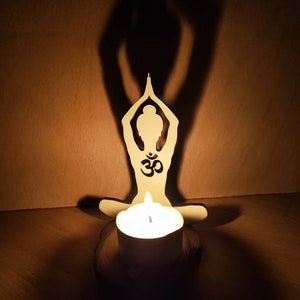 K&ouml;nnte beinhalten: Ein Kerzenhalter aus Holz in Form einer Person in einer Yoga-Pose mit dem Om-Symbol auf der Brust. Der Kerzenhalter ist mit einer wei&szlig;en Kerze beleuchtet.