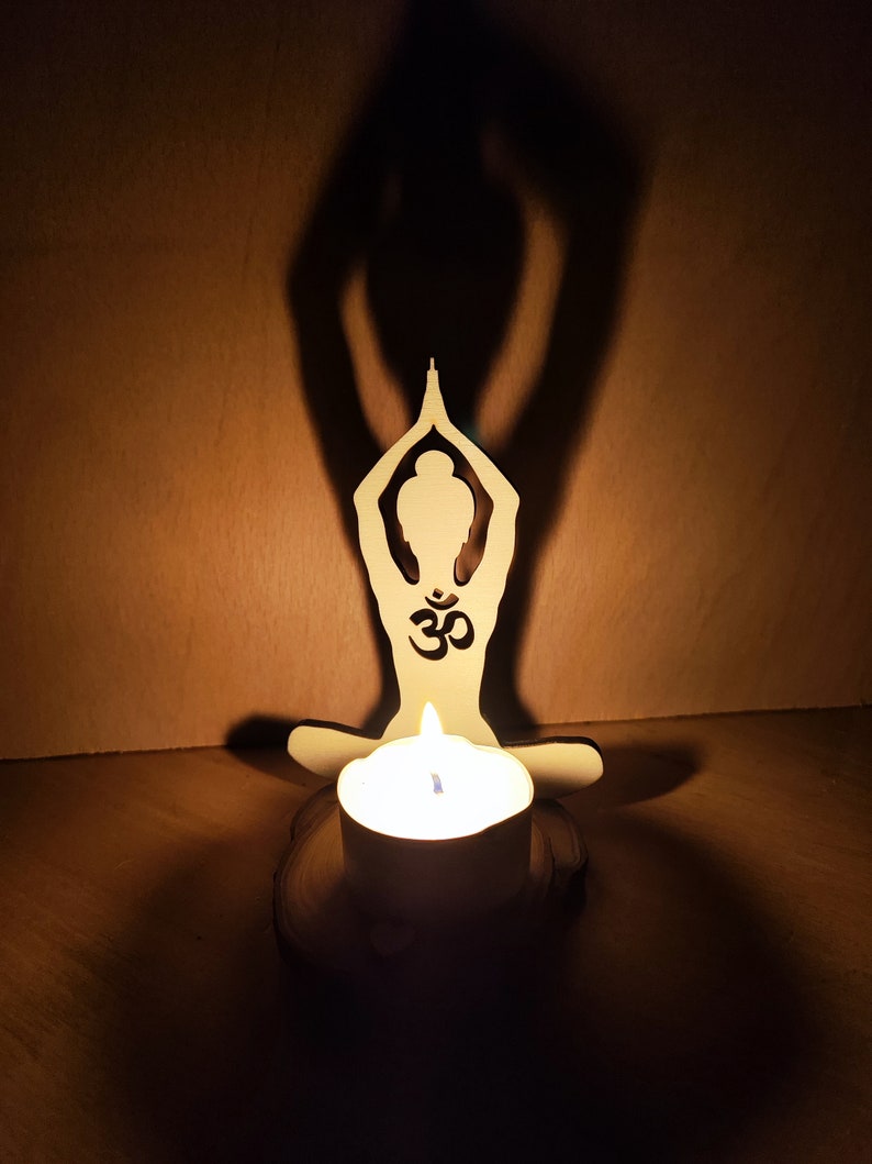 K&ouml;nnte beinhalten: Ein Kerzenhalter aus Holz in Form einer Person in einer Yoga-Pose mit dem Om-Symbol auf der Brust. Der Kerzenhalter ist mit einer wei&szlig;en Kerze beleuchtet.